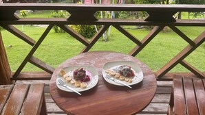 A wooden deck with two round tables, each with a plate of food and a fork. The deck overlooks a lush green landscape with trees and a building in the background. - The Nest Pai Love Resort (Pai)