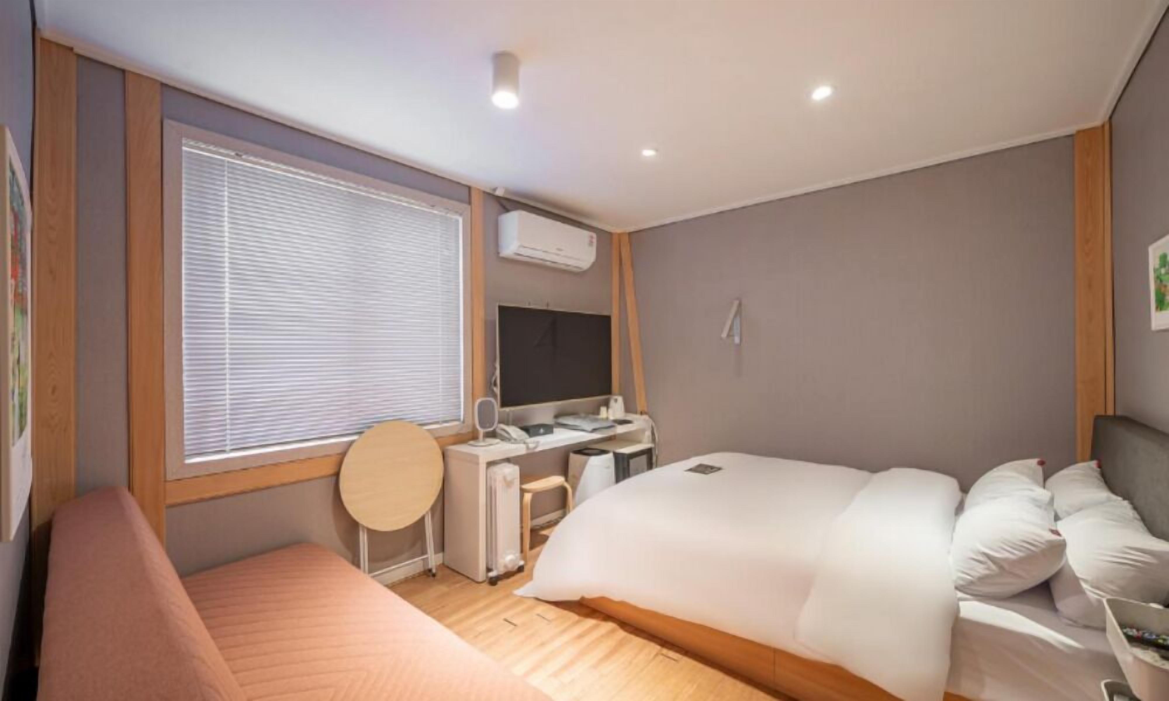 Een slaapkamer met een bed, een bureau, een stoel, een raam met jaloezieën en een airconditioning.