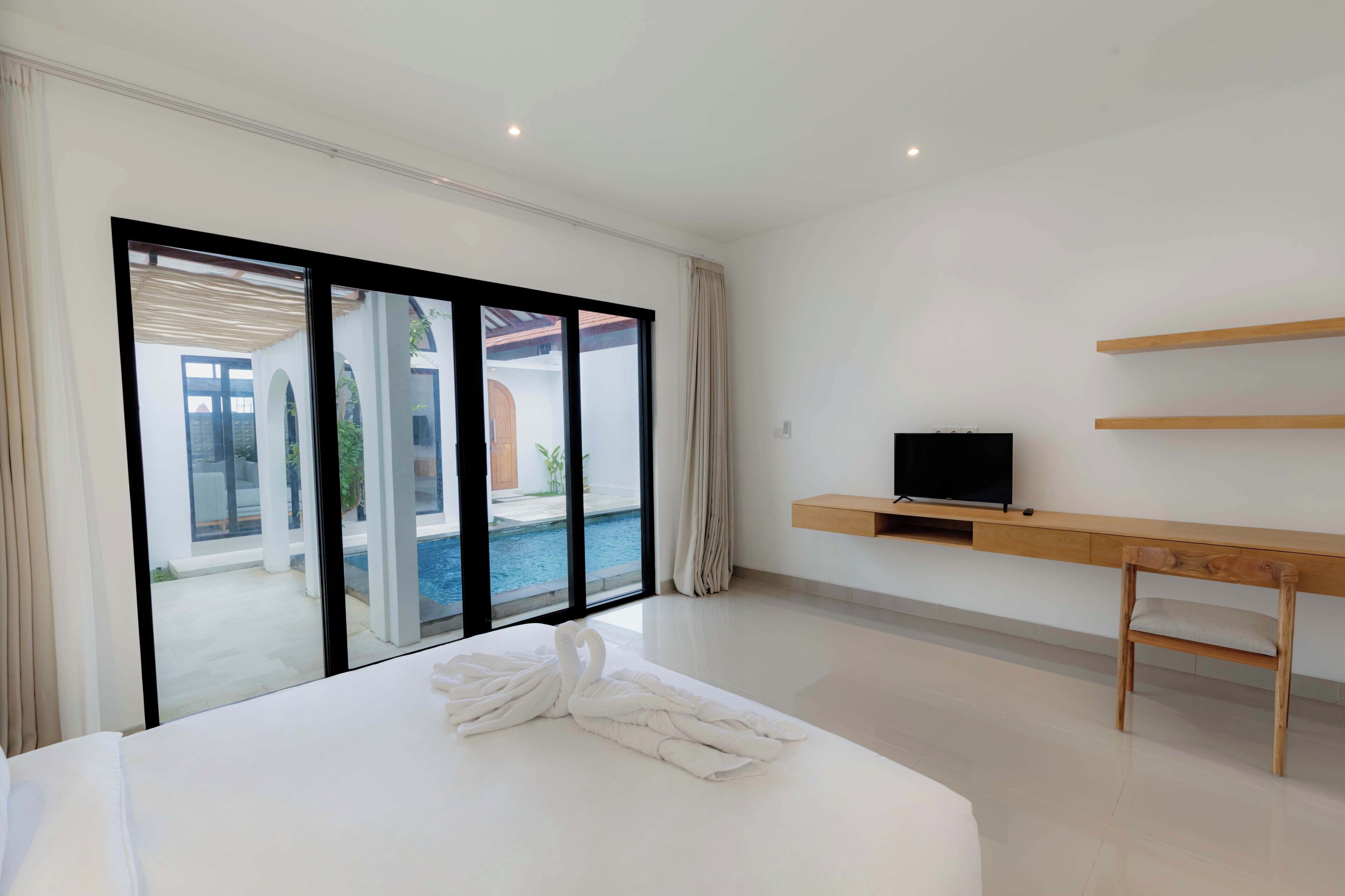 Habitación de hotel moderna con una cama grande, un televisor sobre una consola de madera y vista a una piscina a través de puertas de vidrio amplias.
