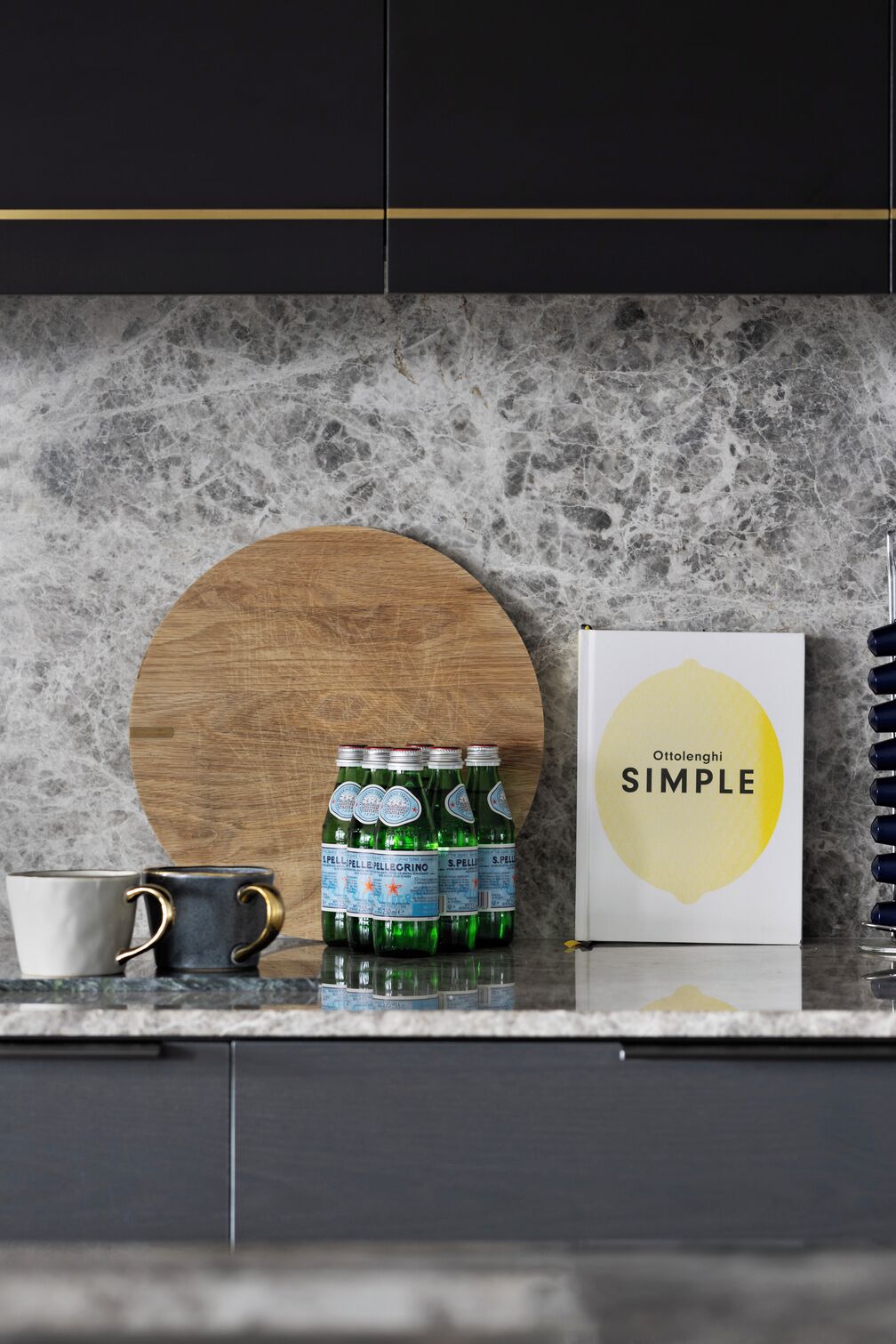 Een keukenblad met een marmeren achterwand, een houten snijplank, een doos met de tekst 'Ottolenghi SIMPLE' en verschillende groene flessen.