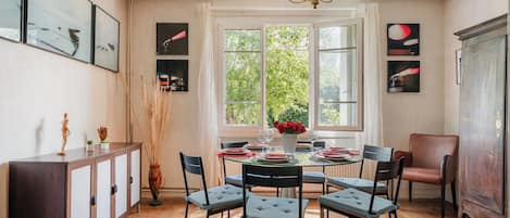 A dining area with a round table, six chairs, a chandelier, and a view of greenery through large windows.