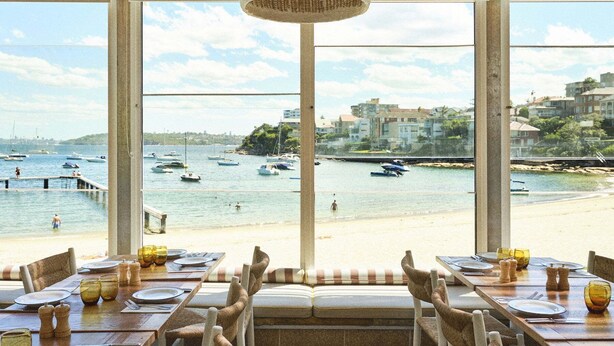 Un espace repas offrant une vue sur la mer et les bateaux, avec des tables en bois, des chaises en osier et un luminaire suspendu.