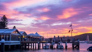Un restaurant situé au bord de l’eau, avec une terrasse, un quai où sont amarrés des bateaux et un ciel au coucher du soleil.