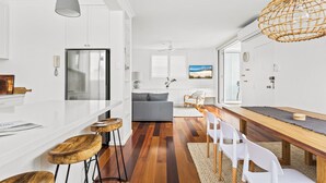 Une cuisine moderne avec des meubles blancs, un grand îlot central doté de tabourets de bar, un coin repas avec une table en bois et des chaises blanches, et un canapé gris en arrière-plan.