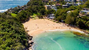 Vue aérienne d’une zone côtière avec une plage, des eaux turquoise et limpides, et une zone résidentielle avec des maisons.
