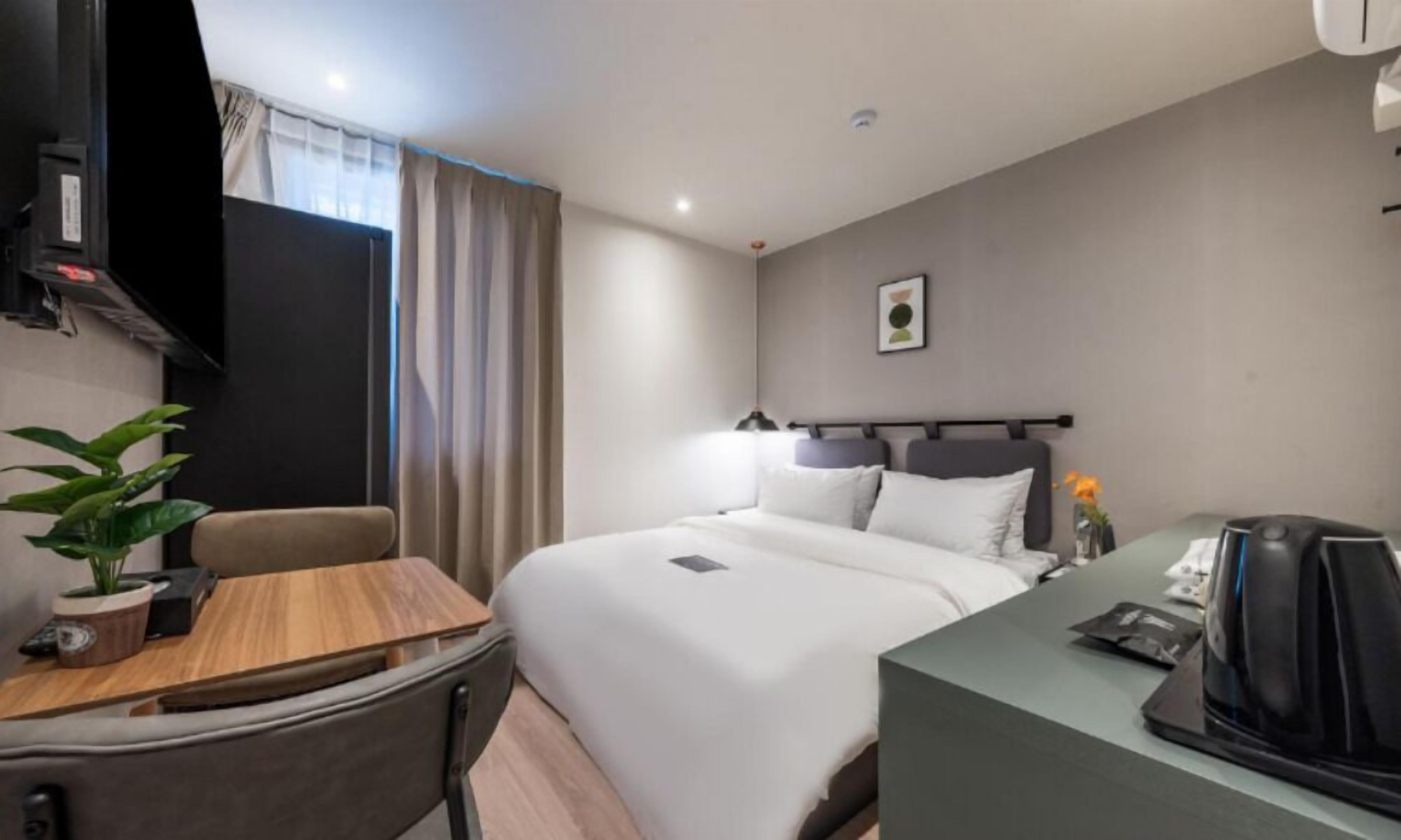 A hotel room with a bed, bedside table, chair, desk, and a potted plant.
