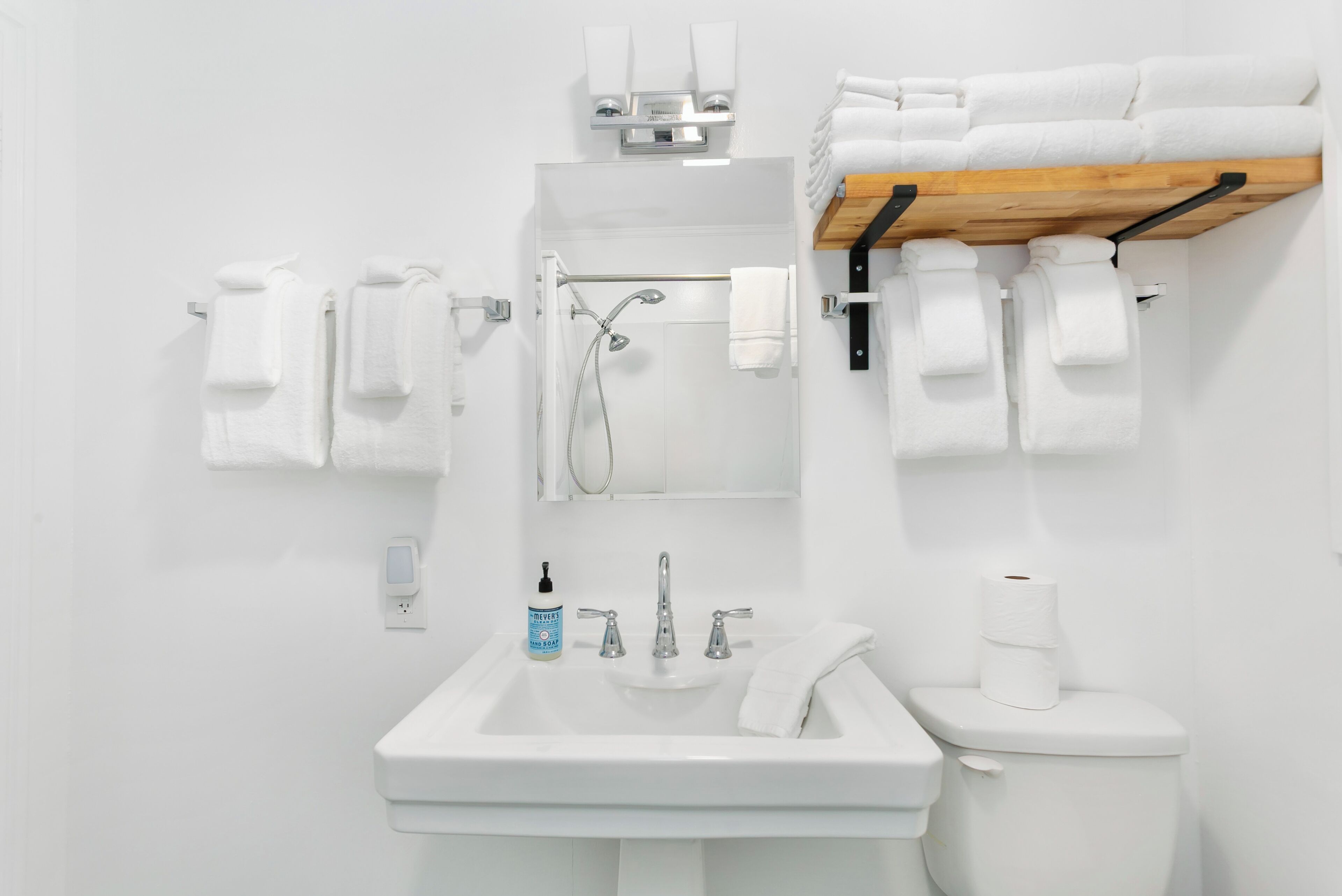 A modern bathroom with a white sink, a toilet, and a shower. There are multiple white towels hanging on hooks and a wooden shelf above the toilet.