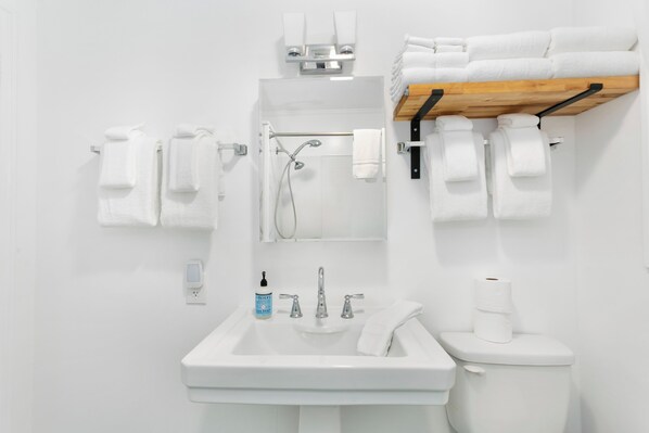 A modern bathroom with a white sink, a toilet, and a shower. There are multiple white towels hanging on hooks and a wooden shelf above the toilet. - The Bee House, Newly Remodeled (Grayland)