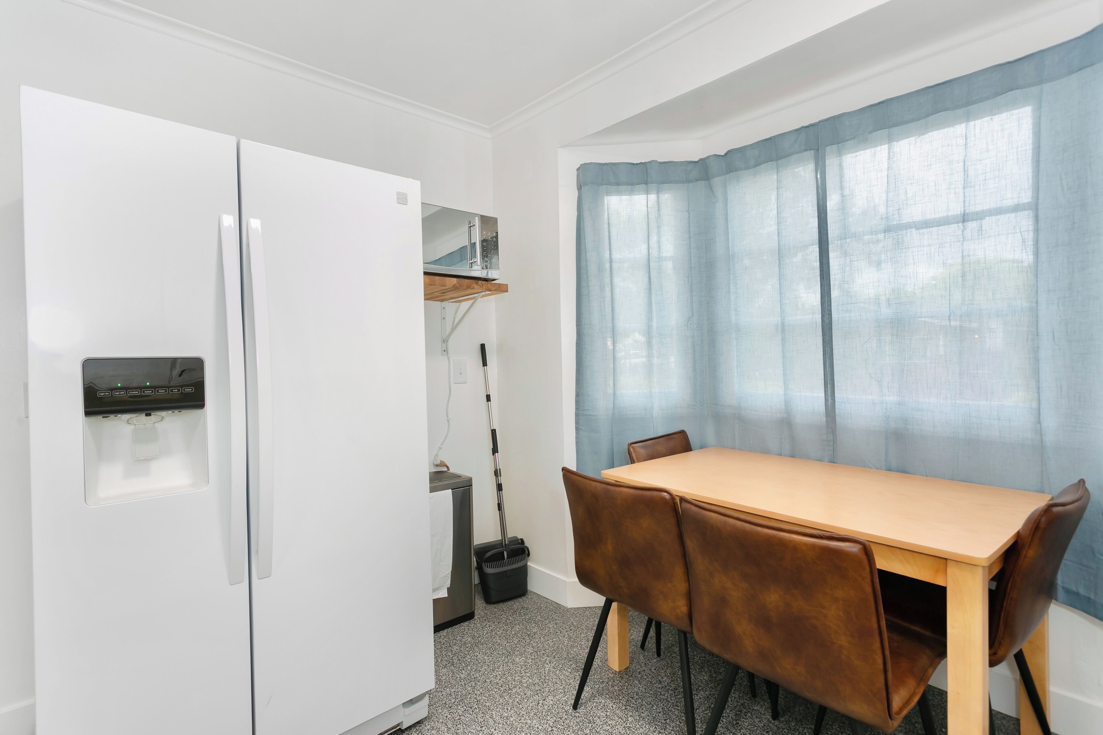 A compact kitchen with a white refrigerator, a small sink, and a broom. There is a wooden table with four chairs and a window with blue curtains.