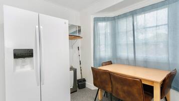 A compact kitchen with a white refrigerator, a small sink, and a broom. There is a wooden table with four chairs and a window with blue curtains.