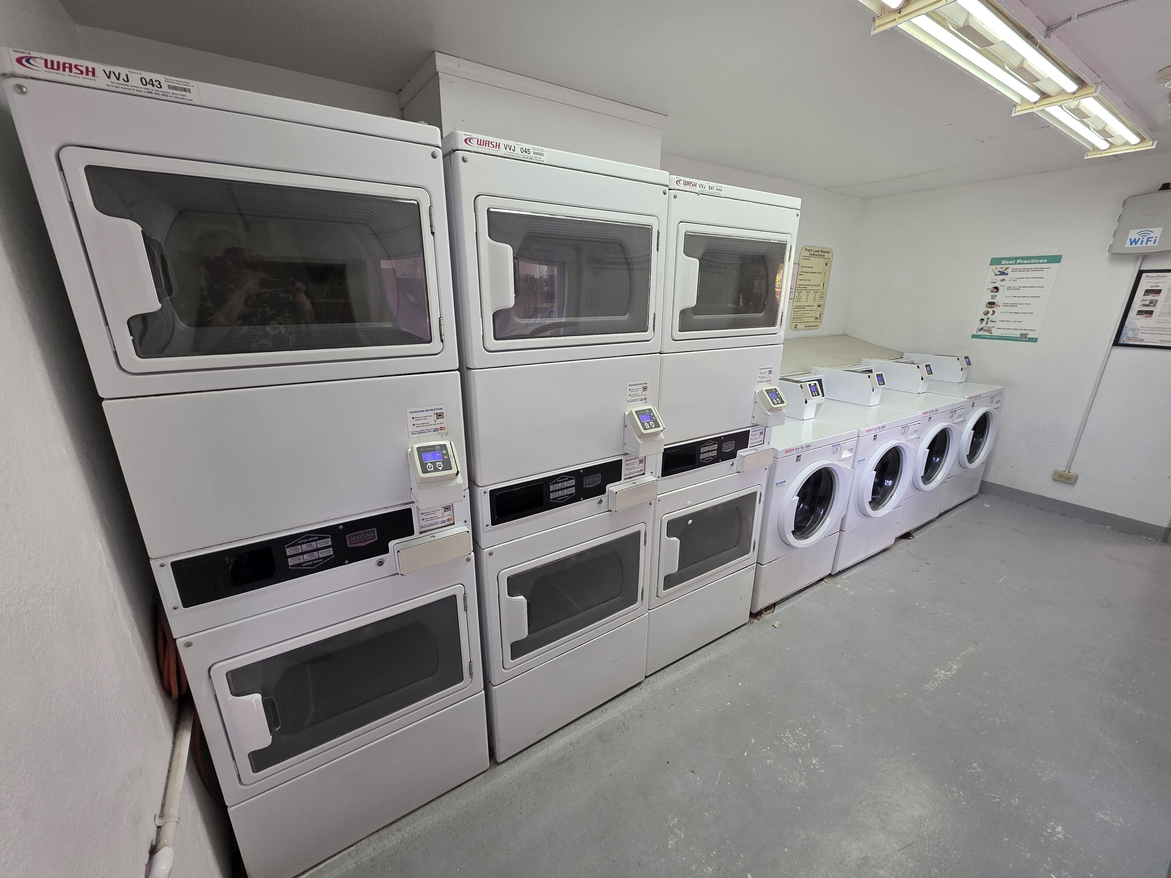 Une laverie équipée de plusieurs machines à laver et de plusieurs sèche-linge, avec un panneau indiquant la présence d’une connexion Wi-Fi et une affiche sur le mur présentant les « meilleures pratiques ».