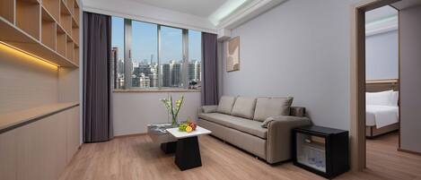 Una habitación de hotel moderna con un ventanal grande, un sofá, una mesita con fruta, un mini refrigerador y una estantería empotrada.