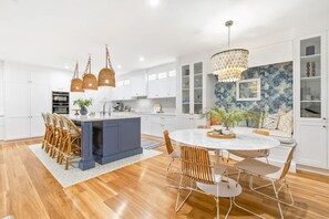 A modern kitchen with white cabinets, a central island, and a dining area with a round table and chairs. - Coastal Luxe (Kingscliff)