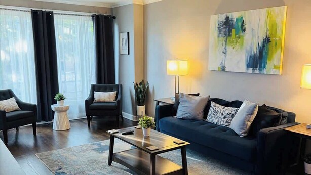 Un salon moderne avec un canapé bleu foncé, deux fauteuils, une table basse et un tapis. On y trouve deux lampes sur des tables d’appoint, un tableau au mur et une fenêtre avec des rideaux.