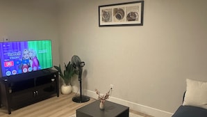 Een woonkamer met een donkergrijze bank, een salontafel, een tv waarop een programma wordt vertoond, een ingelijst kunstwerk aan de muur en een plant in een pot.