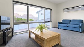 Ein modernes Wohnzimmer mit einem großen Schiebeglastür, einem blauen Sofa, einem Holztisch mit einer Pflanze und einem Flachbildfernseher auf einem Standfuß.