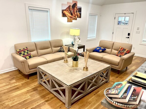 A living room with a beige sectional sofa, a wooden coffee table, and a lamp. There are two windows with blinds, a door, and a wall-mounted picture. - Safari Getaway: MBenz & Beltline (Atlanta)