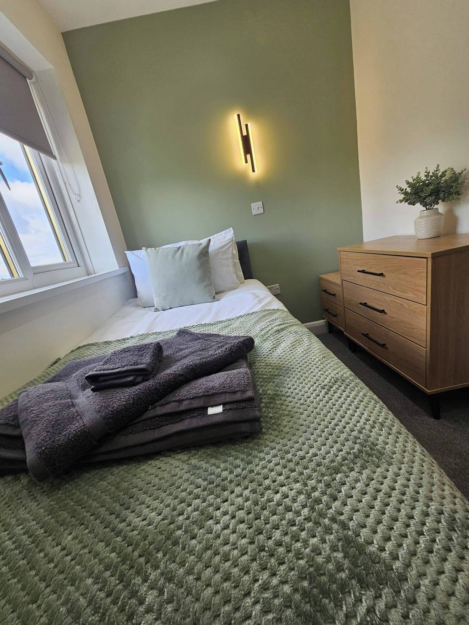 Een keurig opgemaakt bed met een groene sprei, een houten dressoir met lades, en een wandlamp.