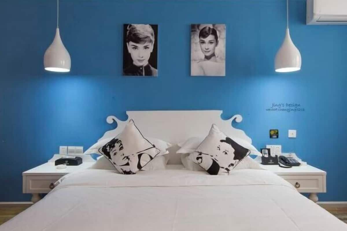 Un dormitorio con cama blanca, dos mesitas de noche blancas, pared azul, dos cuadros enmarcados en blanco y negro, y una lámpara de techo blanca.