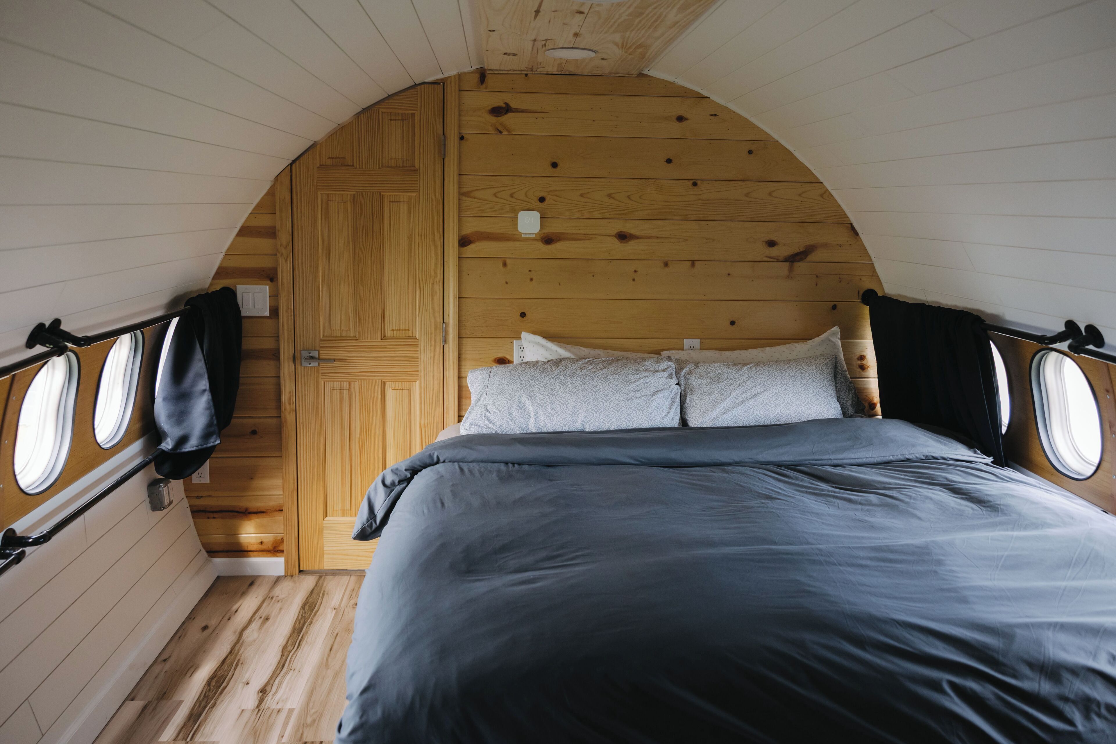 A bedroom with a curved ceiling, wooden walls, a bed with grey bedding, and two windows with curtains.