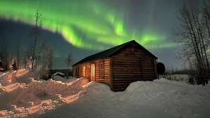 Une cabane en bois, dont le toit et les murs sont recouverts de neige, entourée de congères, sous un ciel nocturne illuminé d’aurores boréales vertes.