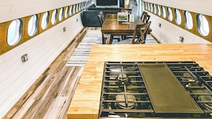 Une pièce étroite et allongée, avec un plafond et des murs en bois, une table centrale sur laquelle se trouve un poêle, et des fenêtres rondes le long des murs latéraux.