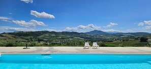 Une piscine avec deux chaises longues blanches, offrant une vue sur des collines verdoyantes et des montagnes sous un ciel dégagé.