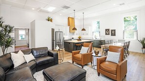 Una sala de estar moderna con un sofá modular, un sillón de cuero marrón y una mesa de centro. Cuenta con una zona de cocina con una isla grande, una mesa de comedor y sillas. La habitación tiene paredes blancas, suelo de madera e iluminación empotrada en el techo.