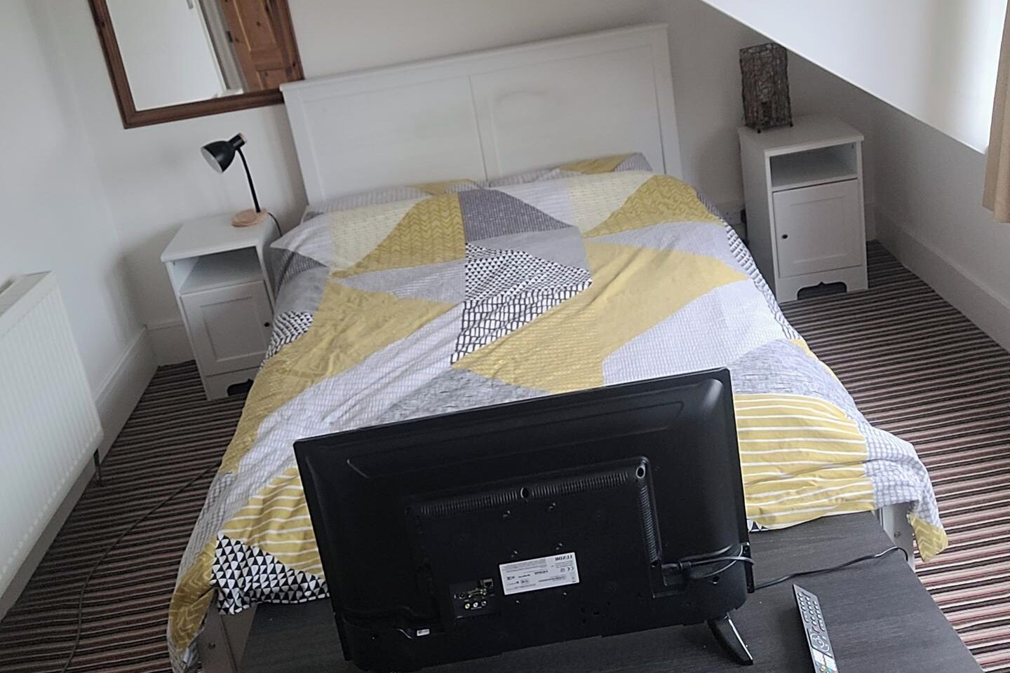 En snyggt bäddad säng med ett geometriskt mönstrat överkast, ett sängbord med en lampa och ett litet sidobord med en dekorativ prydnad.