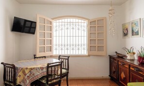 A dining area with a table and chairs, a television mounted on the wall, a decorative window with open shutters, and a cabinet with plants.