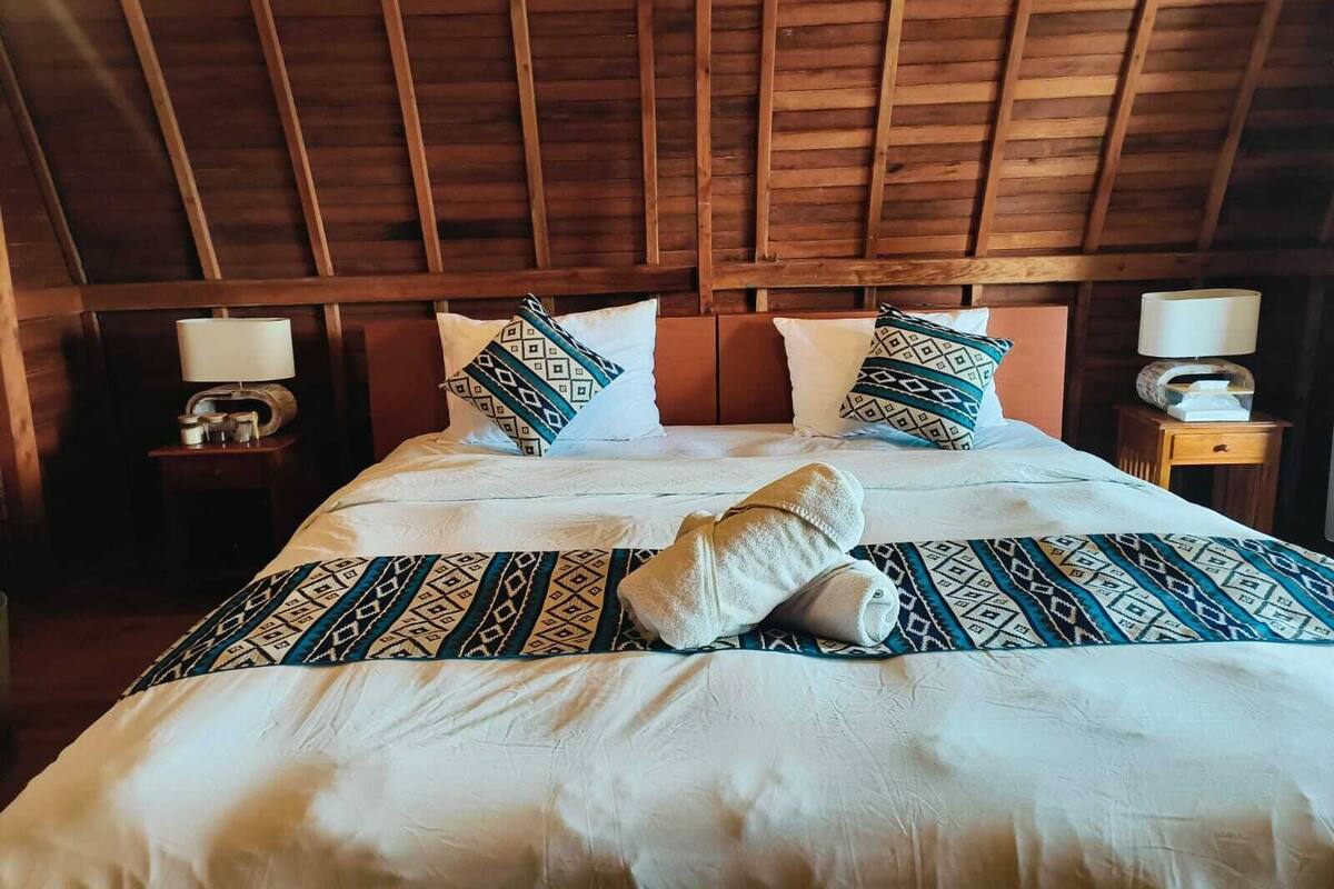 A wooden ceiling in a room with a bed, two bedside tables, and patterned pillows.