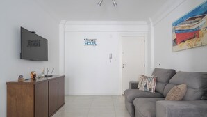 A living room with a grey sofa, a flat-screen TV, a cabinet, and a door with a 'Beach House' sign. - 2-bed Apt w/ BBQ - Downtown Bombinhas Caa103 (Bombinhas)