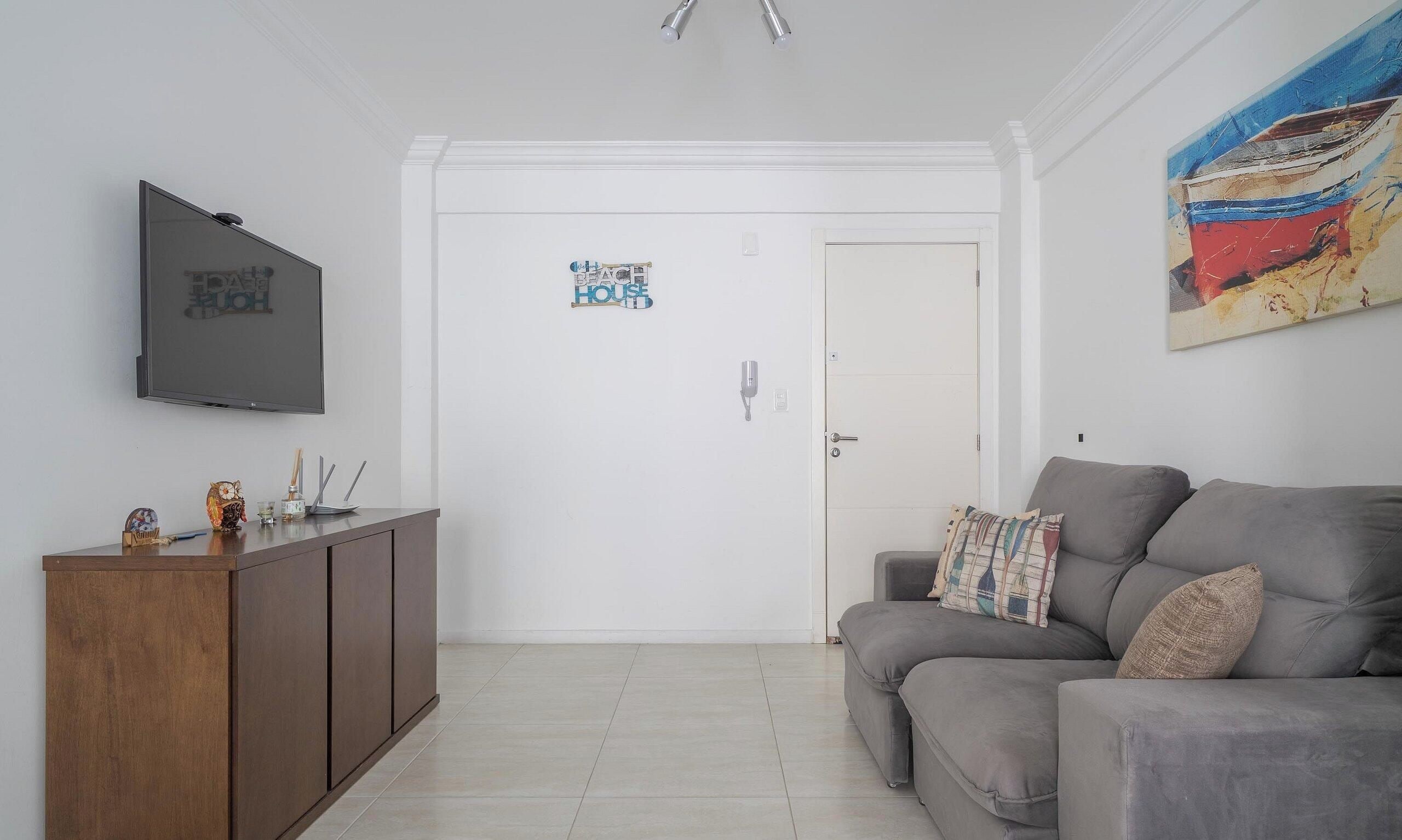 A living room with a flat-screen TV, a wooden cabinet, a grey sofa with patterned cushions, and a door with a 'Beach House' sign.
