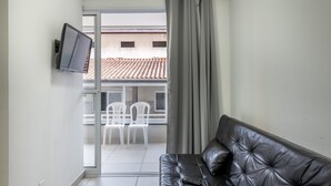 A modern living room with a black leather sofa, a flat-screen TV mounted on the wall, and a sliding glass door leading to an outdoor area with white chairs. - Apt With Barbecue 2 min From Tenorio Beach Cah014 (Ubatuba)