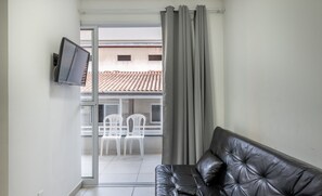 A modern living room with a black leather sofa, a flat-screen TV mounted on the wall, and a sliding glass door leading to an outdoor area with white chairs. - Apt With Barbecue 2 min From Tenorio Beach Cah014 (Ubatuba)