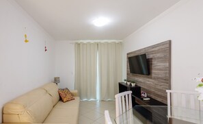 Una sala moderna con un sofá color beige, una mesa de comedor de vidrio, un televisor montado en una pared de madera y una ventana con cortinas blancas.