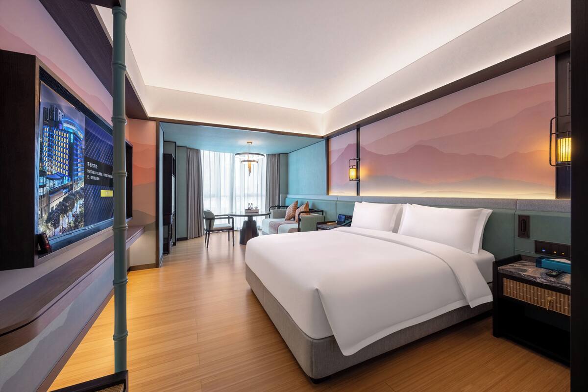 A modern hotel room with a large bed, a desk, a chair, a TV, and a view of a cityscape.