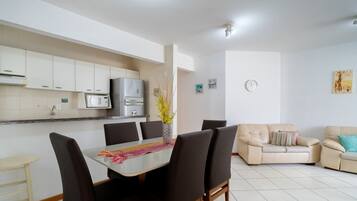 A modern kitchen with white cabinets, a microwave, and a refrigerator. There is a dining table with chairs and a sofa in the living area.