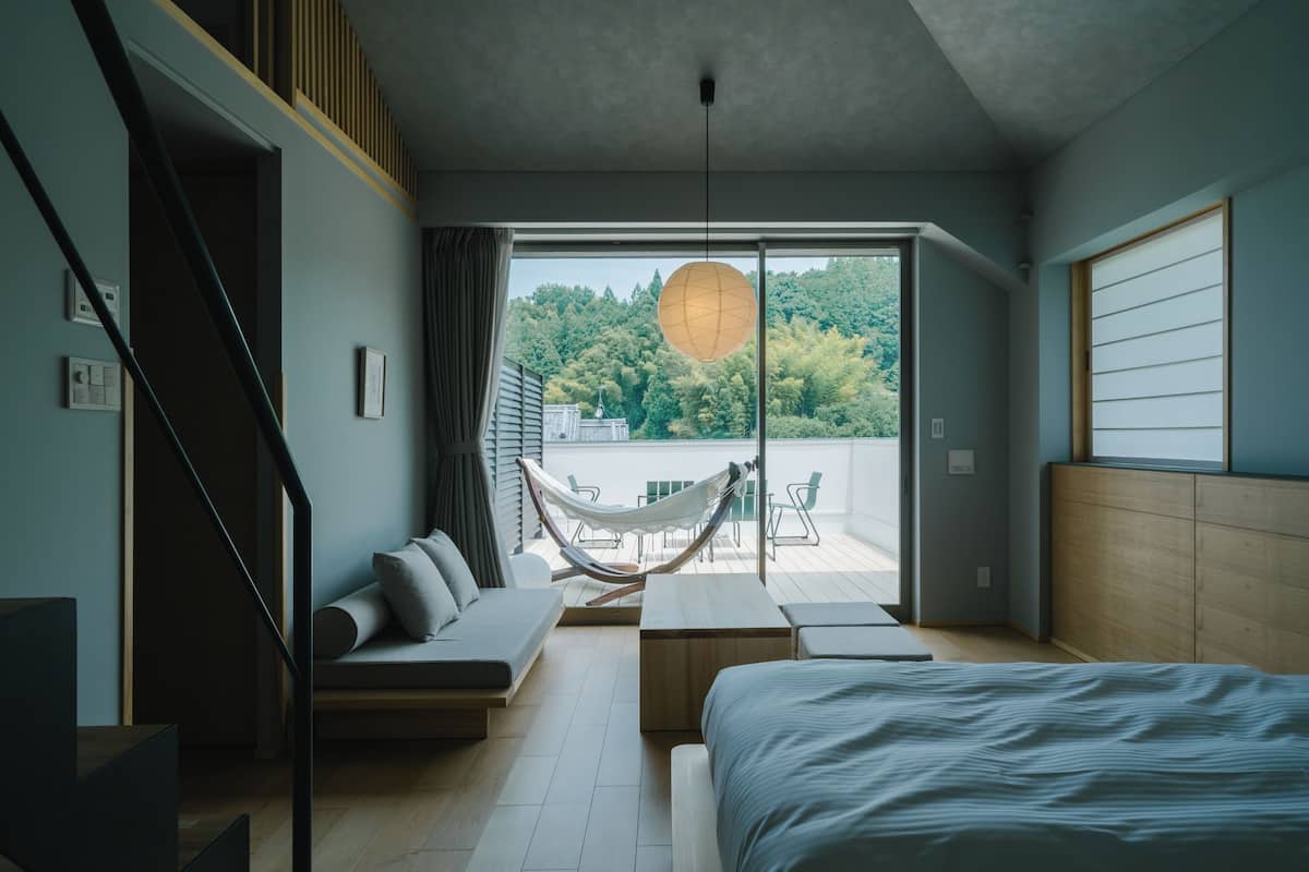 A modern bedroom with a bed, a hammock, a sofa, and a view of a balcony with a wooden railing.