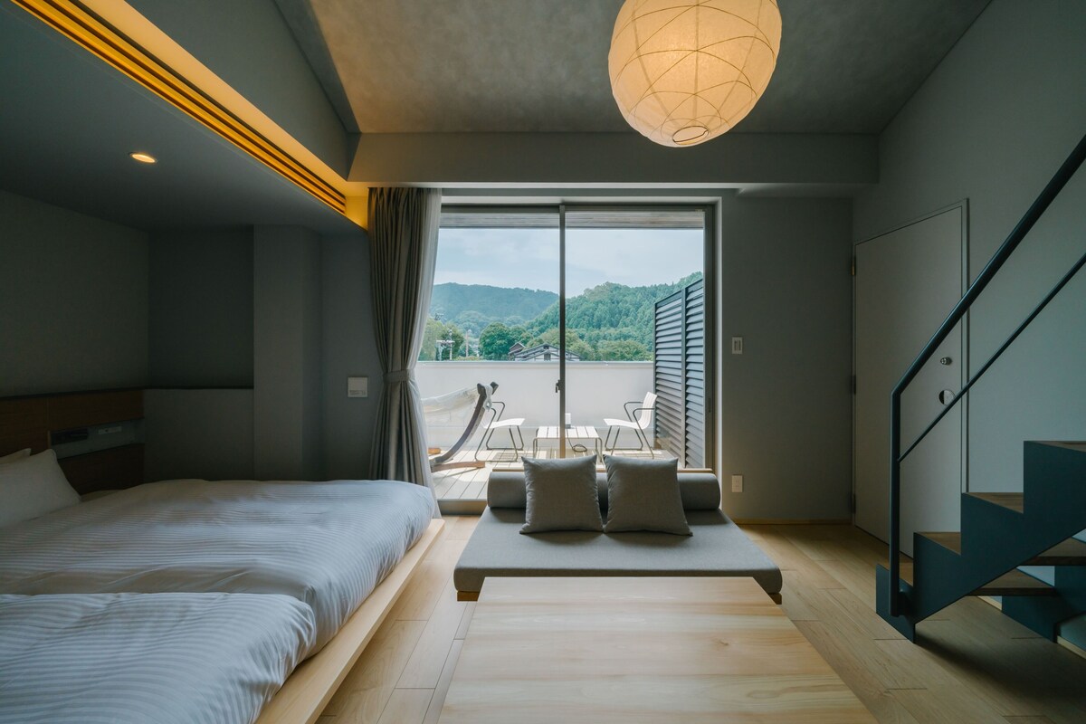 A modern bedroom with a bed, a bench with pillows, a staircase, and a view of a balcony with outdoor furniture.