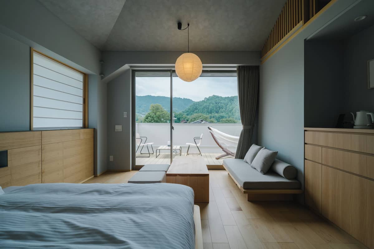 A modern bedroom with a bed, a sofa, a desk, and a view of a balcony with a hammock.