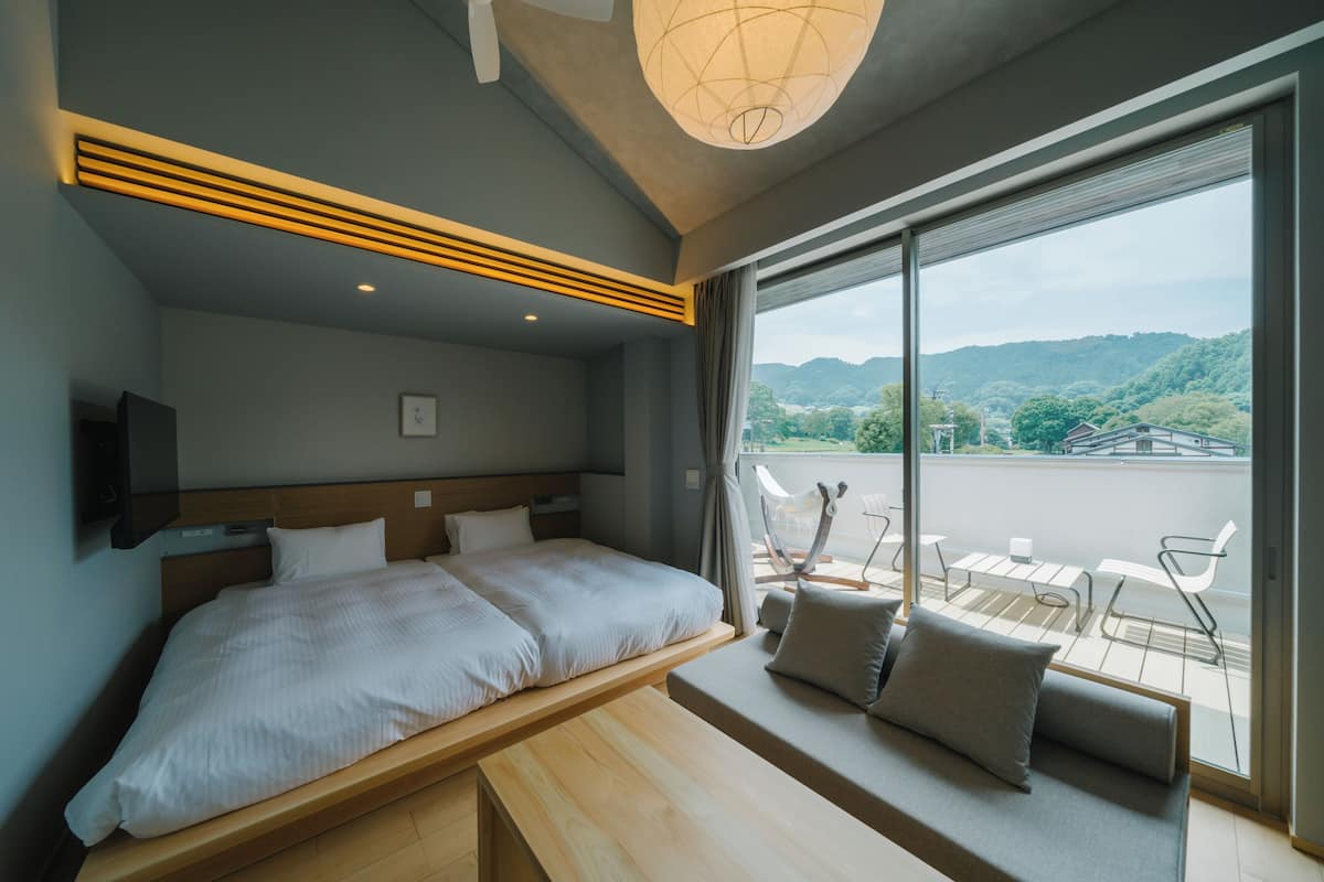 A modern hotel room with a large bed, a wooden bench, and a balcony with a view of the countryside.