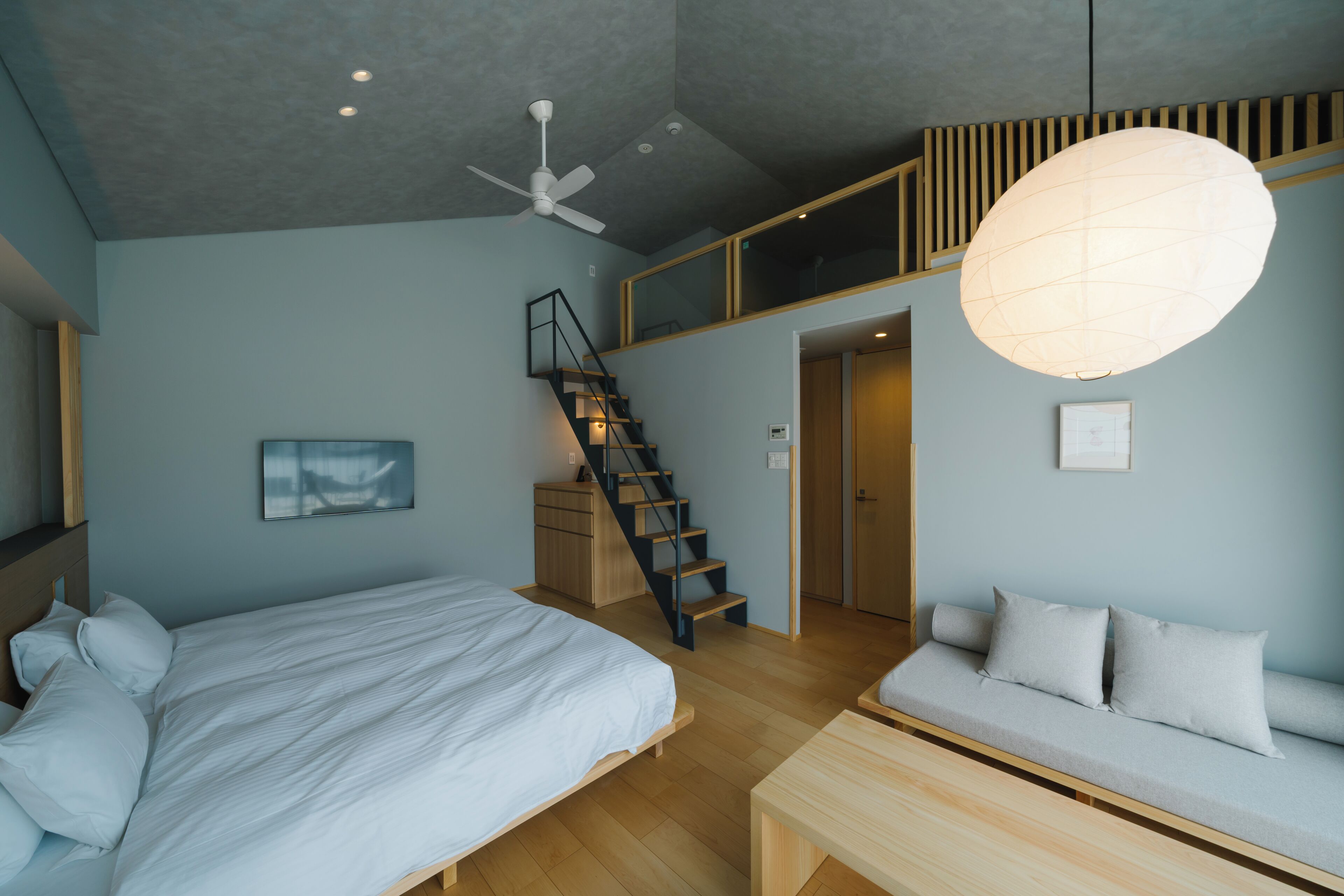 A bedroom with a bed, a sofa, a desk, a staircase, and a ceiling fan.