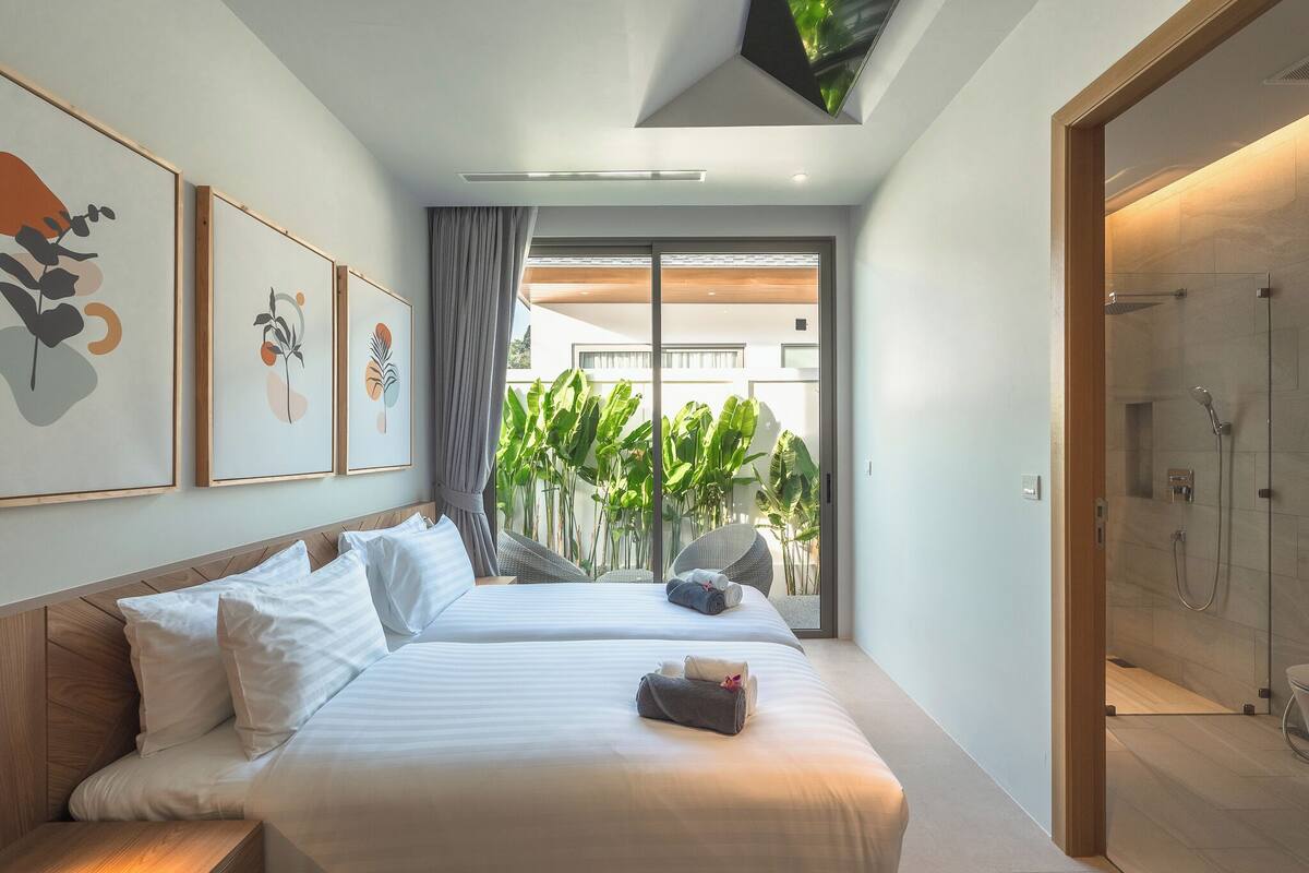 A modern hotel room with two beds, a shower, and a view of greenery outside.