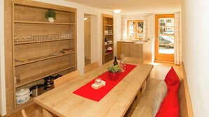 A compact kitchen with wooden shelves, a dining table with a red placemat, and a view of the street through an open door. - Apartment Typ C by Interhome (Längenfeld)