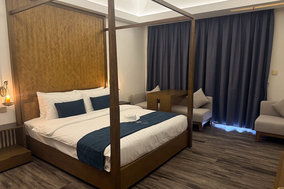 Una cama con dosel y tablero de madera, una mesita de noche, un sofá y una silla en una habitación de hotel con un papel tapiz de dibujo de espiga y cortinas oscuras.