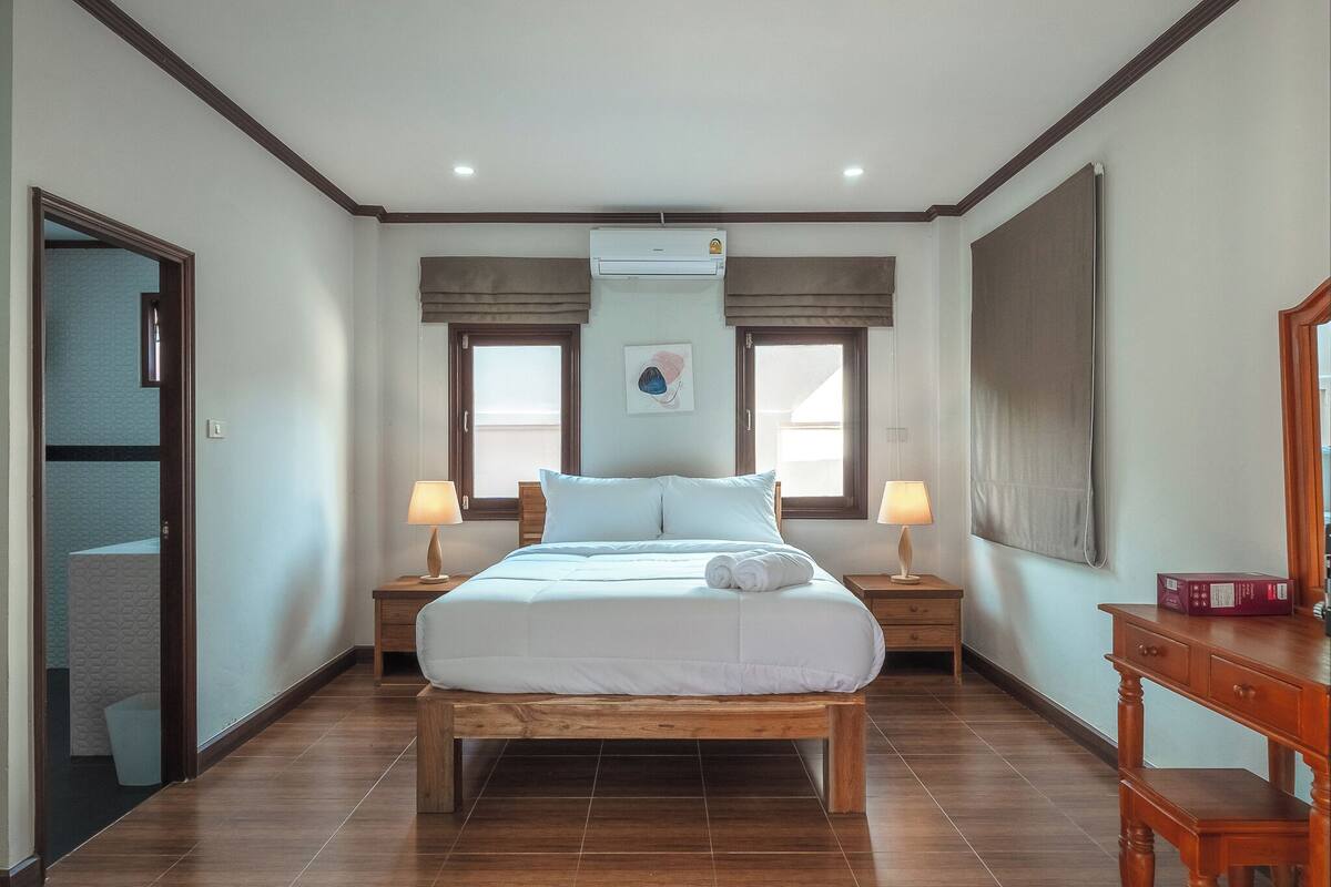 A bedroom with a wooden bed, two bedside tables with lamps, a large window with curtains, and a wall-mounted air conditioner.