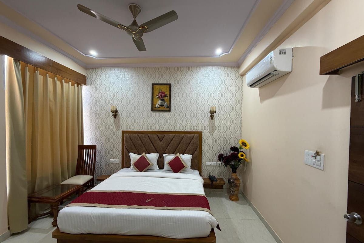 Une chambre d’hôtel comprenant un lit, un ventilateur de plafond, un appareil de climatisation, une table de chevet, une chaise et un vase avec des fleurs.