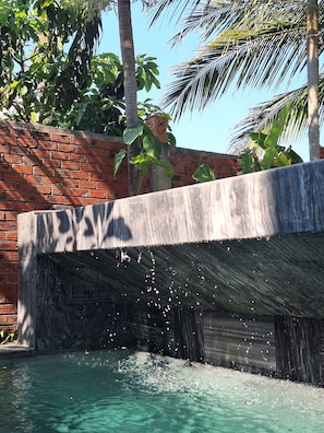 Un élément décoratif aquatique en béton, agrémenté d’une cascade, entouré d’un mur de briques et de palmiers.