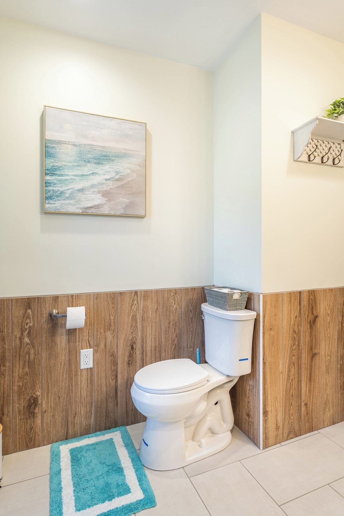 Une salle de bain avec un mur recouvert de panneaux en bois, des toilettes blanches, un tapis bleu et un tableau représentant une scène de plage.